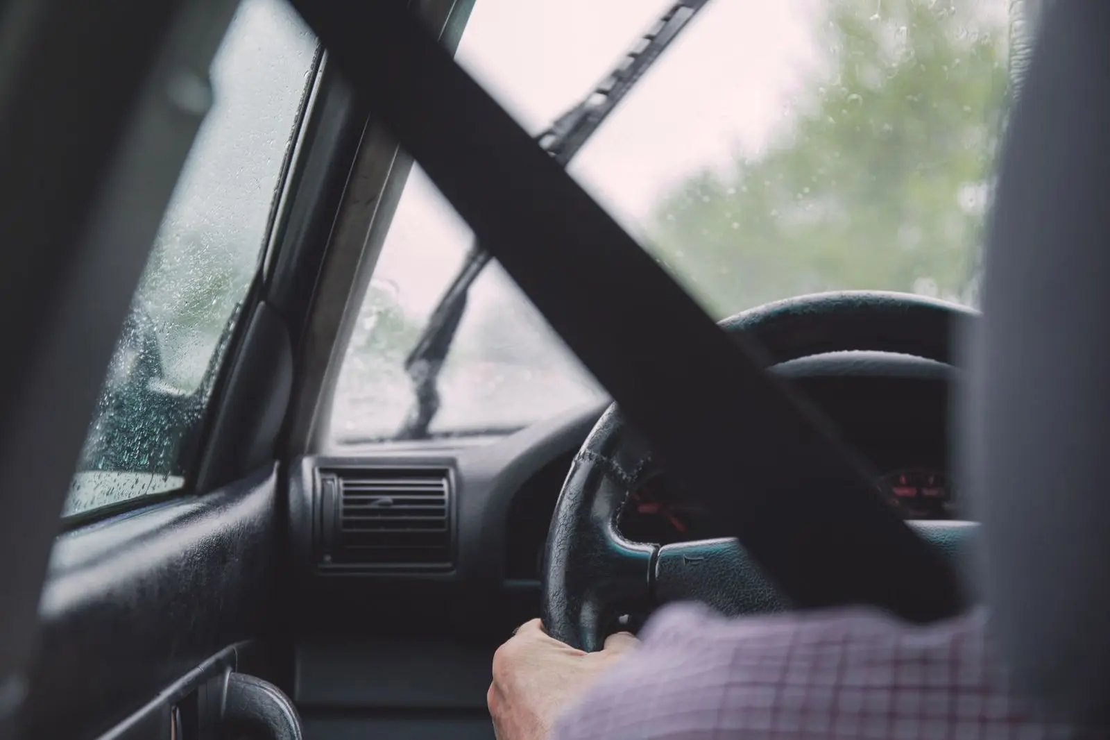 A driver at the wheel in rain, seatbelt across the chest, wipers on the windscreen.