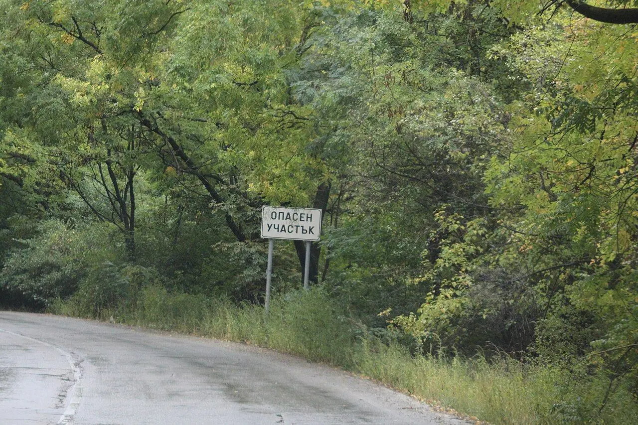 A white rectangular sign in Bulgarian reading dangerous section, beside a narrow wet road.
