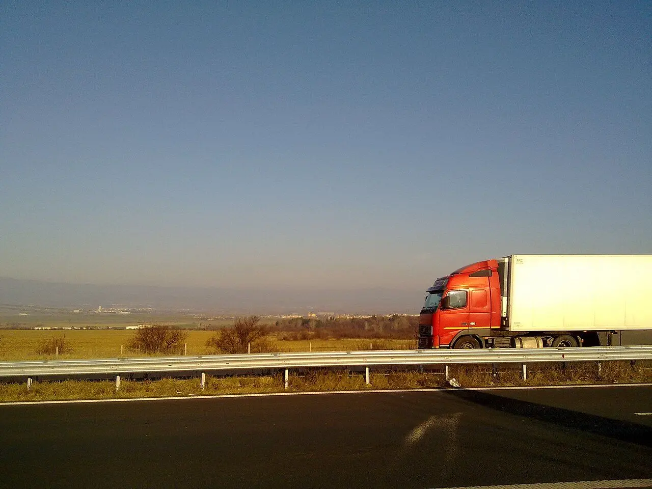 A red tractor unit with a white trailer on the Trakia motorway, fields beyond the guardrail.
